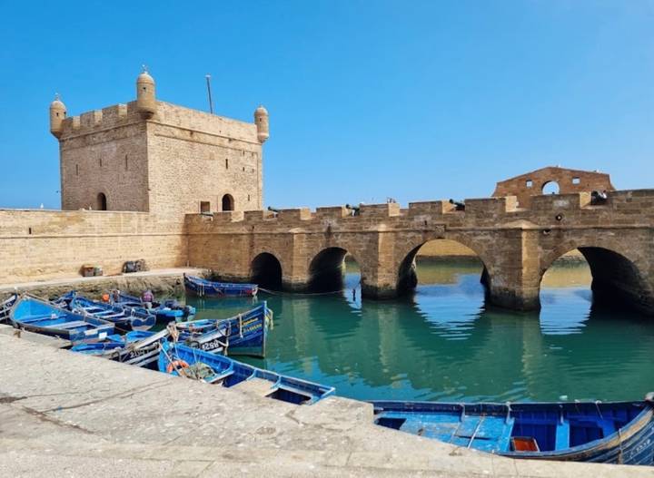 An old stone fort and arched bridge guard a calm green inlet dotted with blue fishing skiffs.