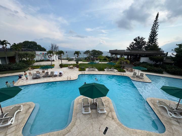 A resort pool overlooks misty Lake Atitlán and distant volcano peaks under a cloudy sky.