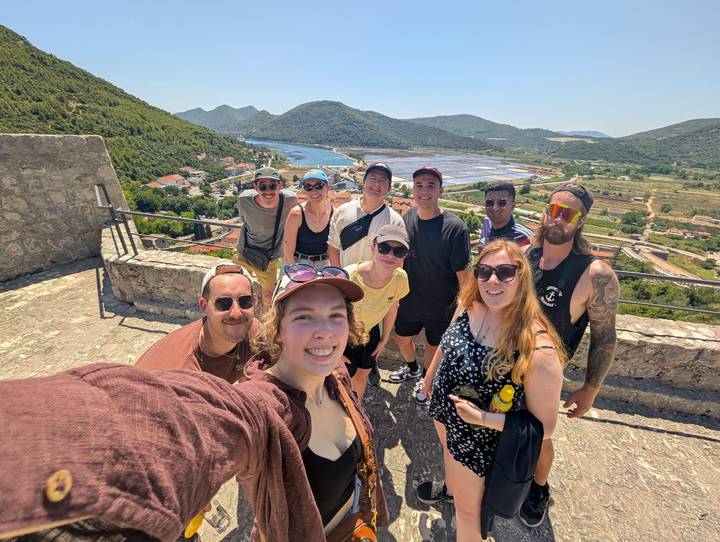 A group of smiling travellers taking a selfie at a hilltop lookout with salt pans, river and green hills in the background.