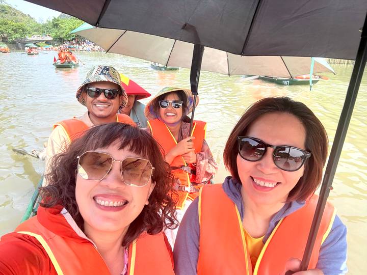 Friends in orange life-jackets smiling on a small rowboat, holding an umbrella on a muddy river.