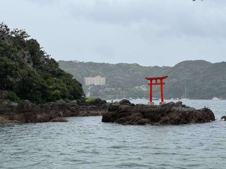 Small red torii gate standing on rocky islet in coastal bay surrounded by forested hills