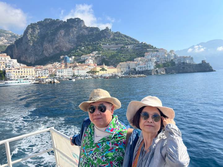Smiling couple on boat with Amalfi Coast village and cliffs in background on sunny day.