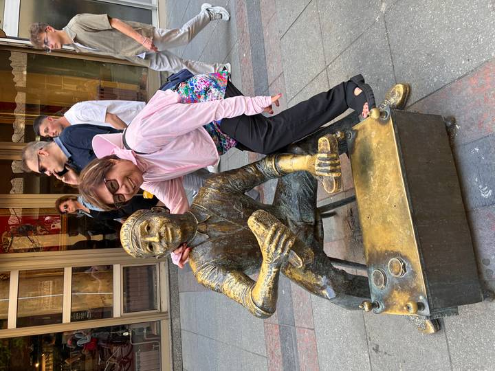 Woman playfully posing with bronze shoeshine statue on pedestrian street.