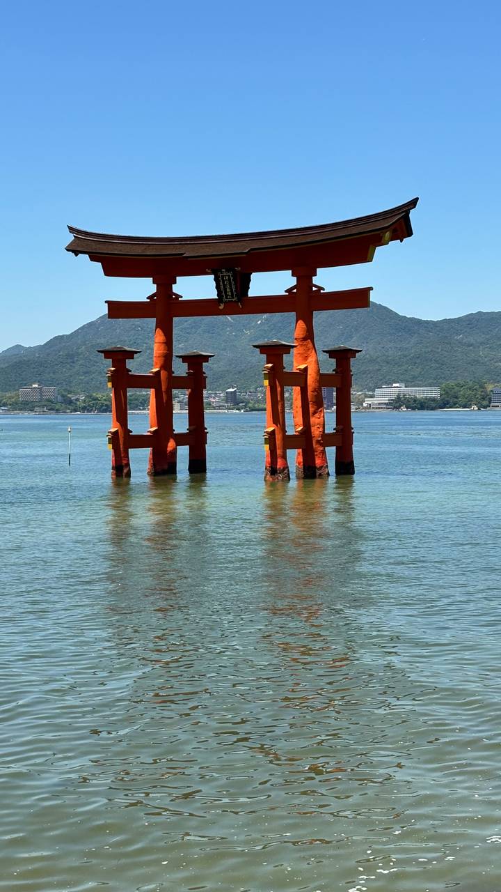 Iconic orange torii gate of Itsukushima Shrine standing in calm bay waters with mountains beyond.