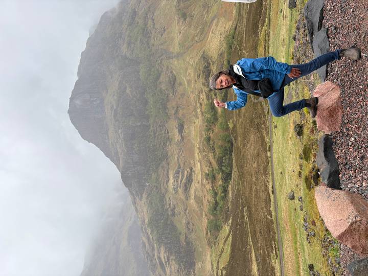 A woman in a blue jacket triumphantly poses on a rock with misty Scottish mountains behind.