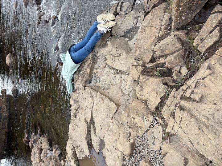 Person lying face-down on riverside rocks reaching into the clear stream.