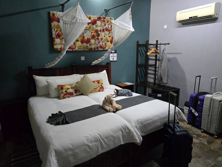 Neat twin-bed lodge room with mosquito nets, suitcases and folded towel animal.