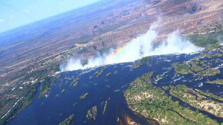 Oblique aerial of Victoria Falls with rising mist and a vivid rainbow cutting across the canyon.