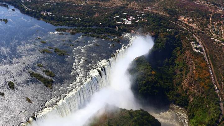 High aerial showing Victoria Falls curtain of water plunging into gorge surrounded by forest.