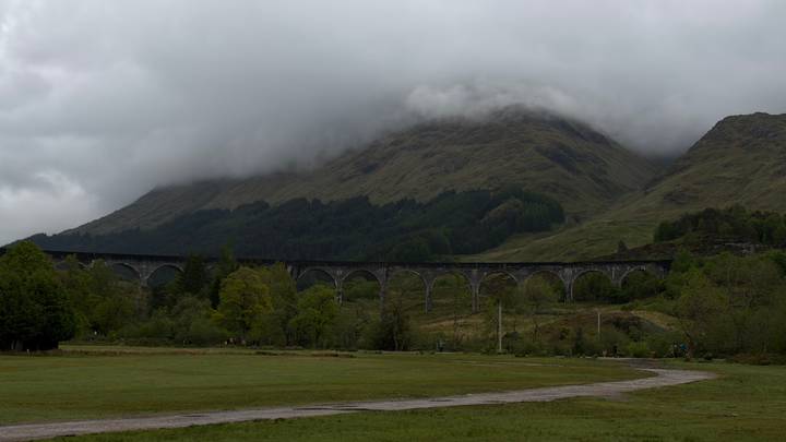 The Glenfinnan Viaduct arcs across a green valley beneath mist-shrouded highland mountains and low gray clouds.