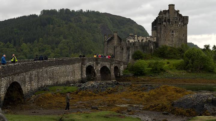 Eilean Donan Castle stands on a small island linked by an old stone bridge, framed by rugged hills and tidal shore at low tide.