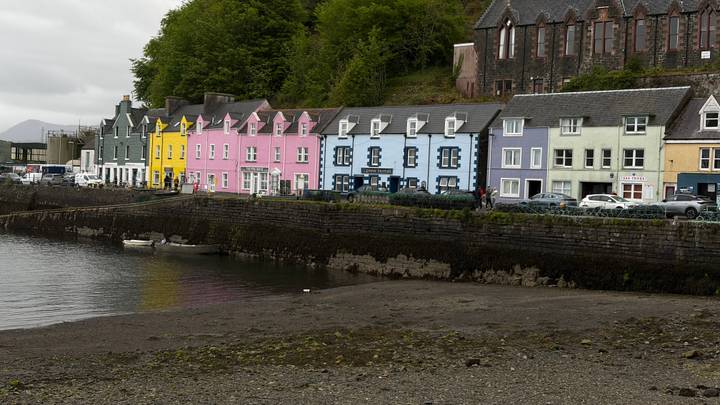A row of pastel-coloured terraced houses lines the harbour front of Portree beside calm water and a rocky shore.