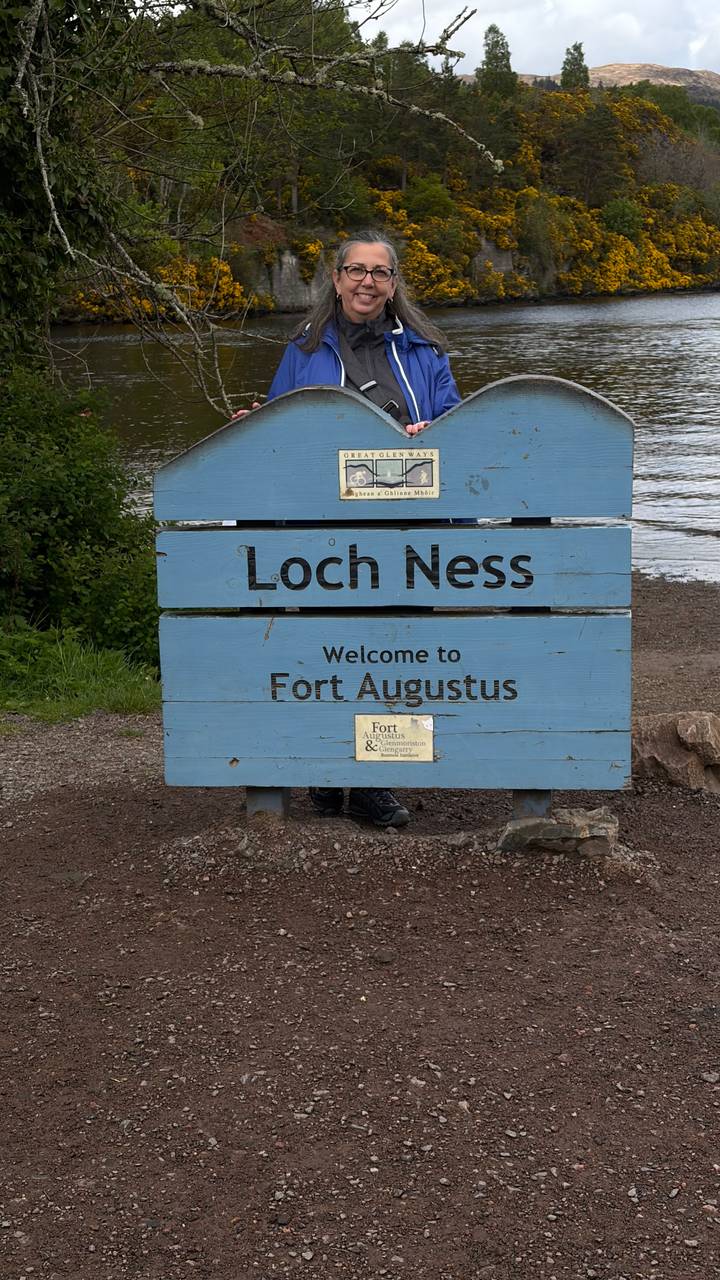 A blue wooden sign reads "Loch Ness – Welcome to Fort Augustus" by the lakeshore.