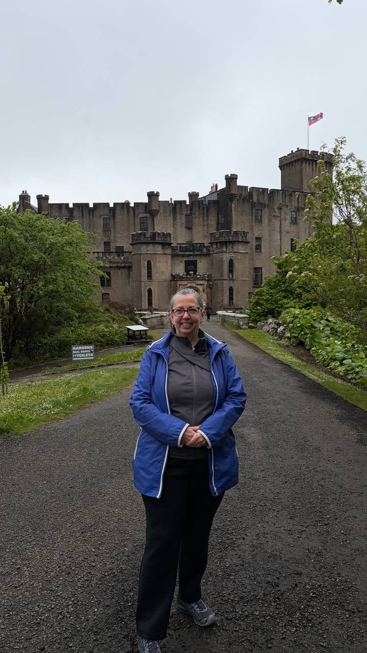 A smiling traveller in a blue rain jacket poses on a driveway leading to a historic Scottish castle surrounded by lush gardens.