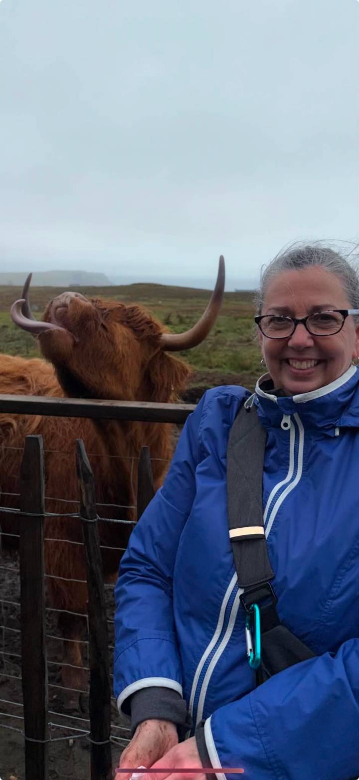 Smiling woman posing near friendly Highland cow stretching its head over a wooden fence
