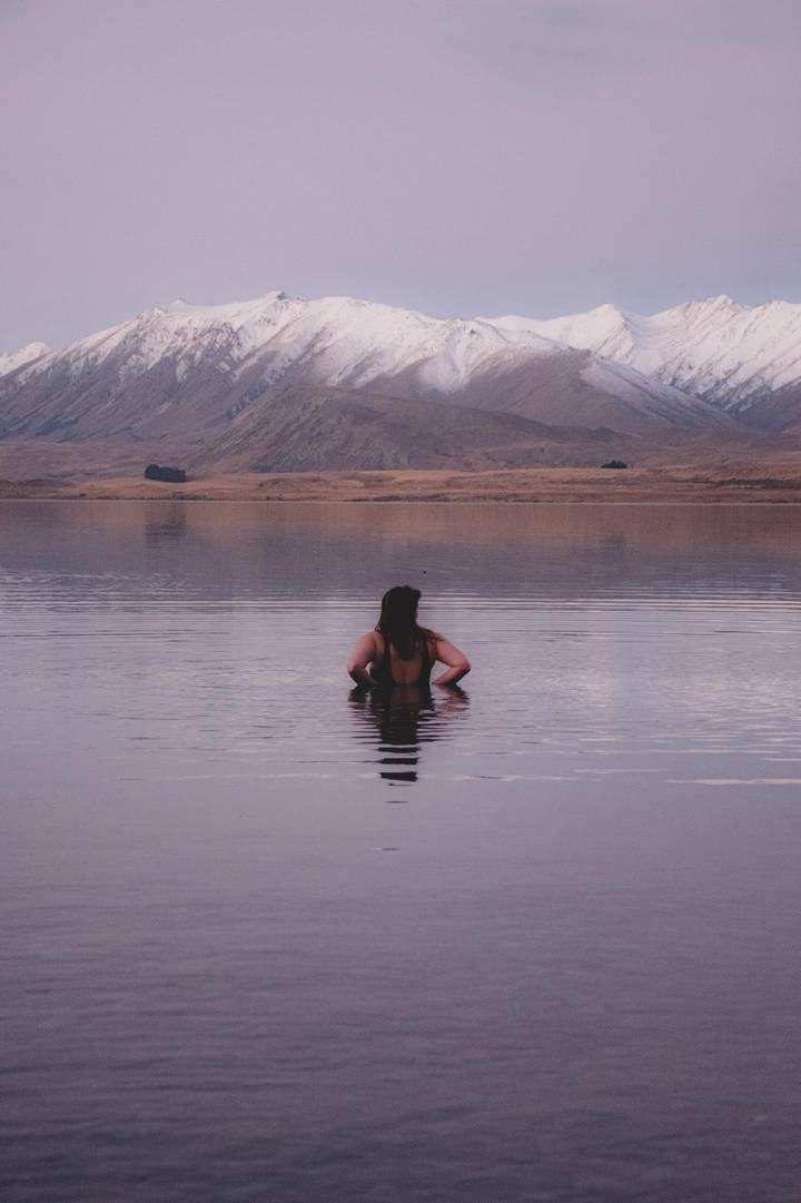 Woman standing waist-deep in tranquil lake at dusk with mountains reflecting on calm water