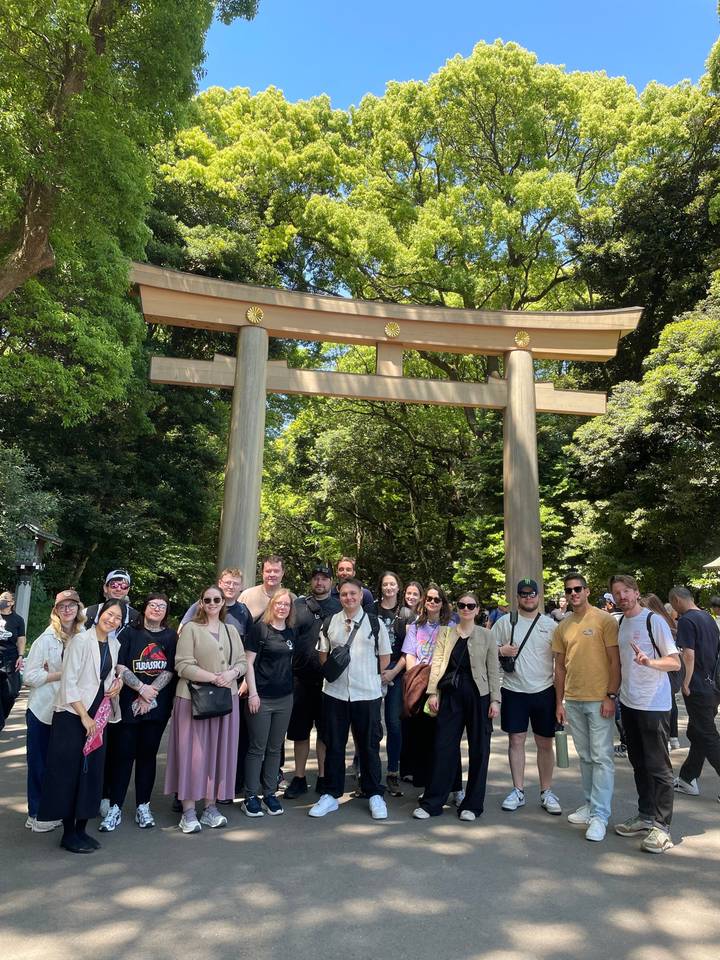 Large tour group posing below a wooden torii gate surrounded by lush forest in bright sunlight.