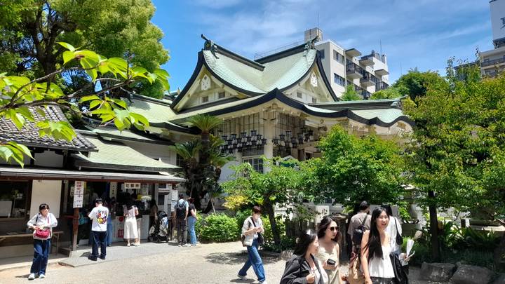 Busy Shinto shrine courtyard filled with greenery, visitors and nearby city apartment blocks.