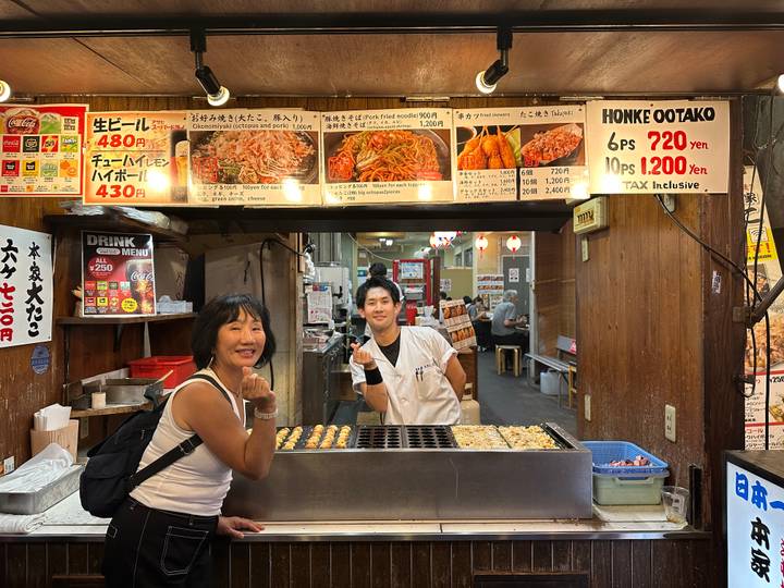 Customer smiles at a bustling food stall as the chef prepares takoyaki behind the griddle.