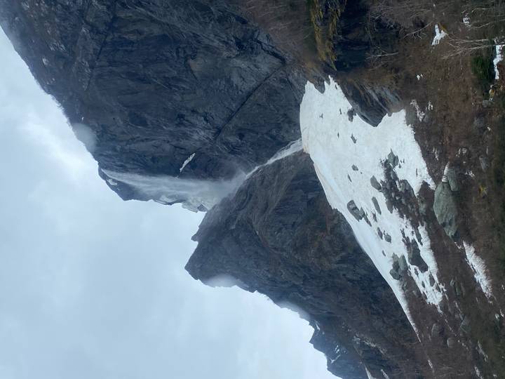 Tall waterfall plunges down a sheer cliff beside lingering spring snow and misty skies.