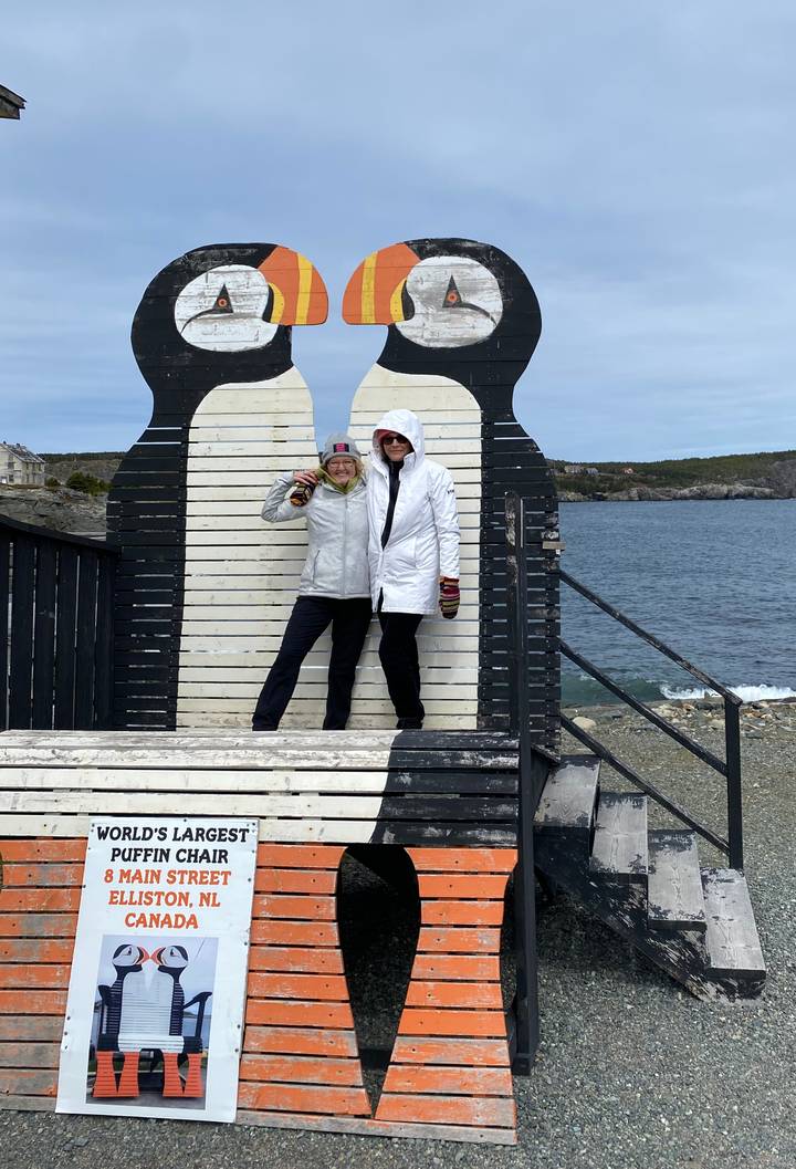 Two friends in coats pose inside a giant black-and-white wooden chair by the sea.