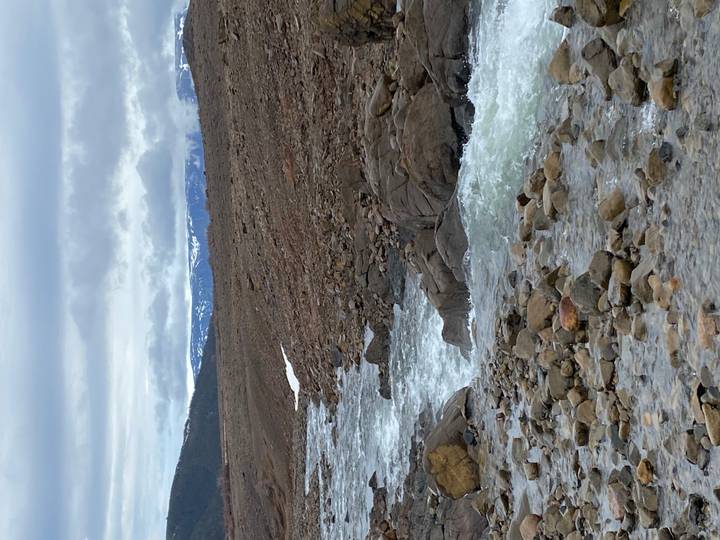 Fast-flowing rocky river cuts through a barren tundra with distant snowy mountains under grey skies.