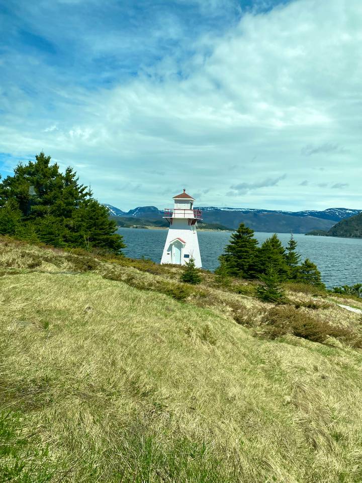 A small white and red lighthouse stands on a grassy hill overlooking a calm lake and distant snow-capped mountains beneath a partly cloudy sky.