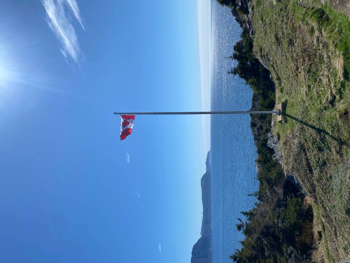 The Canadian flag waves from a tall pole above a rugged green coastline and sparkling blue ocean on a bright clear day.