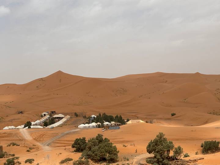 White desert camp tents cluster at the base of vast orange sand dunes under a hazy sky.