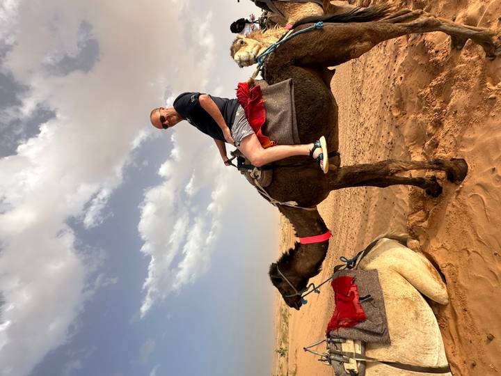 A smiling traveler rides a camel across reddish desert sands beneath scattered clouds.