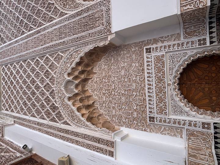 Intricately carved plaster and geometric patterns frame an ornate arched doorway in a Moroccan madrasa.