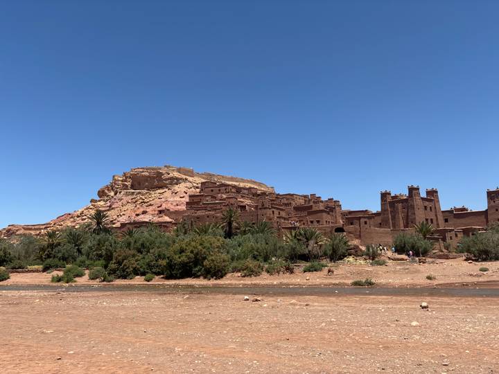 The fortified clay ksar of Aït Benhaddou crowns a hill above palm trees and a shallow river under a clear blue sky.