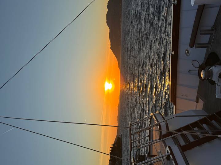 Golden sun setting over rippling sea viewed from the deck of a yacht with distant silhouetted hills.