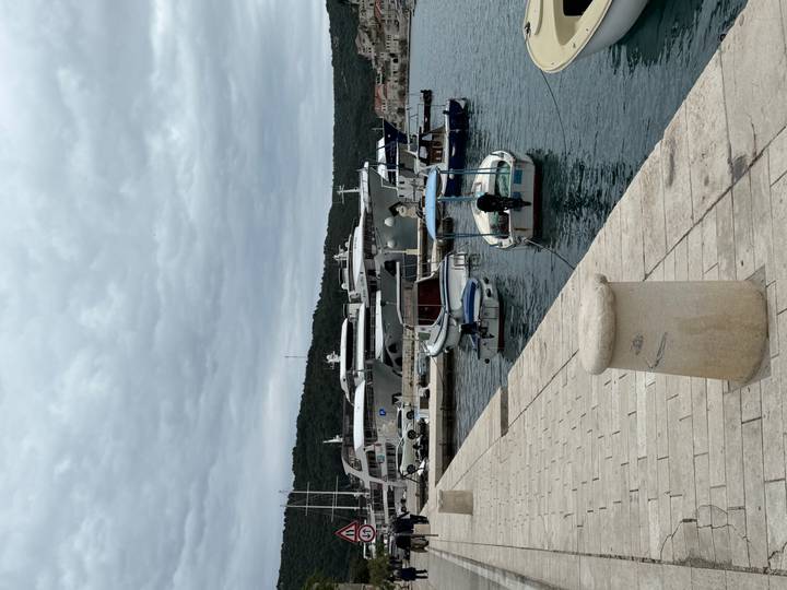 Small marina with moored yachts and tenders under a cloudy sky, stone quay in the foreground.