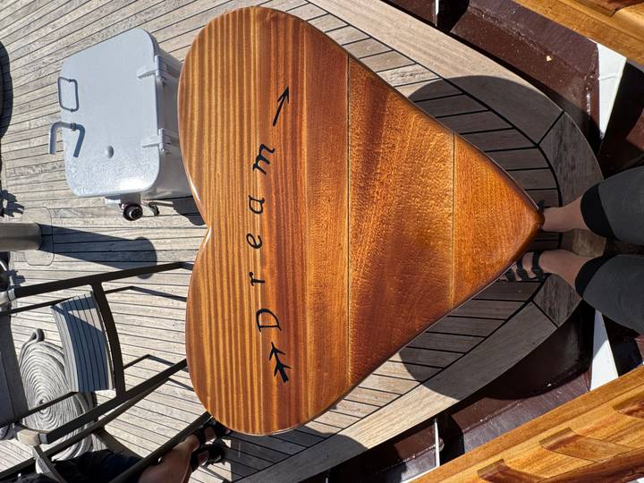 Heart-shaped polished wooden table inscribed "Dream" on a teak deck with a traveler's legs visible below.
