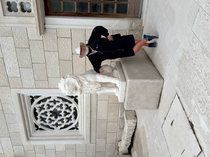 Older traveler poses beside a carved stone lion outside an ornate limestone building.