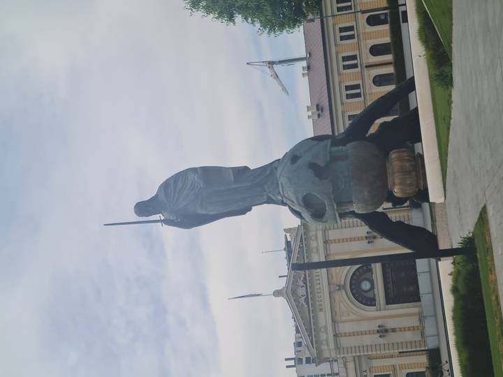 Modern monumental statue of a cloaked figure standing atop a globe outside a historic building.