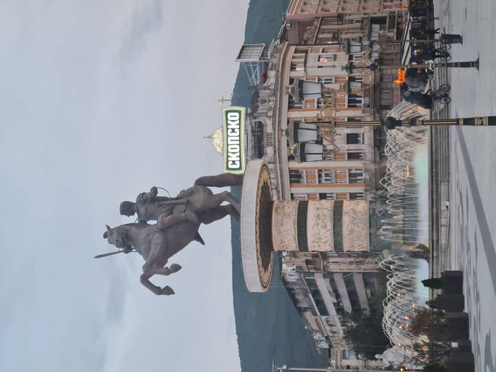 Equestrian statue fountains and neon signage dominate a busy city square at dusk in Skopje.