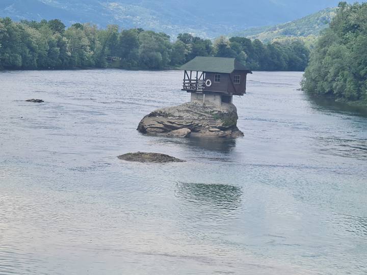 Tiny wooden house balanced on a solitary rock in the middle of a wide river surrounded by forest.