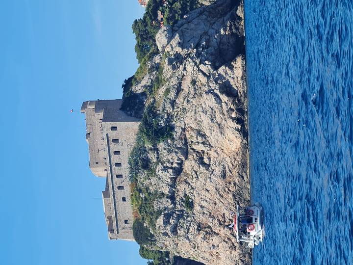 Stone coastal fortress rises from a rocky cliff above the vivid blue Adriatic Sea with a tour boat nearby.