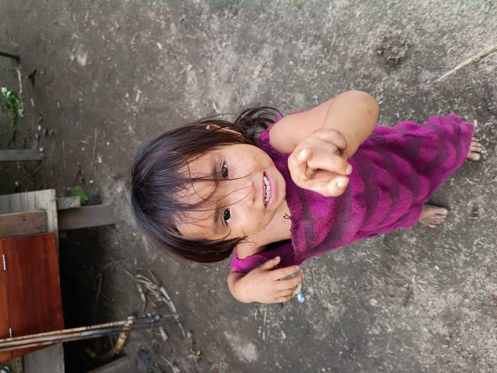 Smiling barefoot child wrapped in a purple towel pointing towards the camera on a dirt yard.