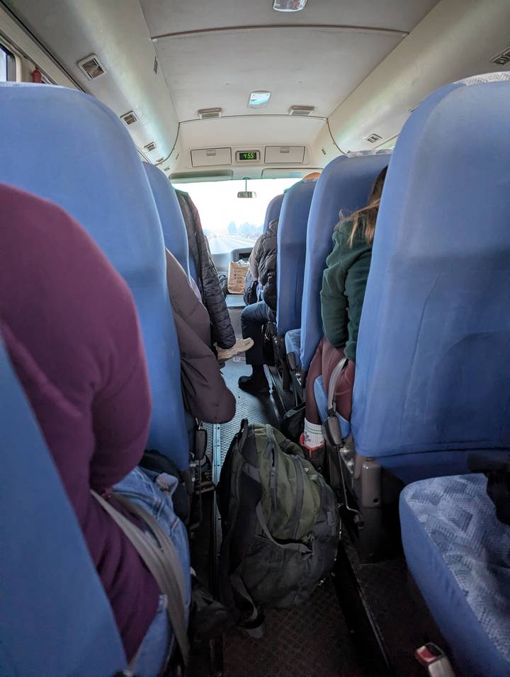 View down the aisle of a tour bus with passengers seated in blue fabric seats.