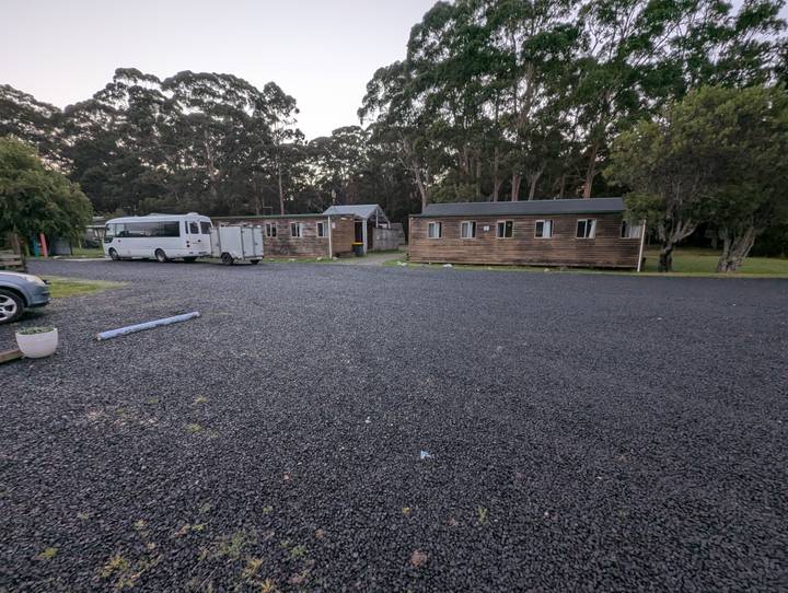 Wide gravel lot with wooden cabins, trailer and camper van under tall trees.