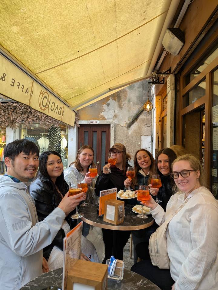 Seven young travelers raise Aperol spritz cocktails and smile inside a cozy Italian bar with rustic walls.
