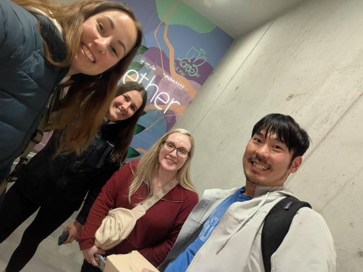 Four friends take a casual indoor selfie in front of a colourful poster that reads "together."