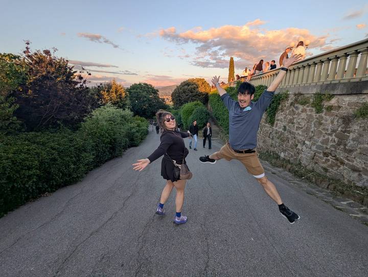 Two travellers joyfully jumping on a hillside lane with sunset clouds and a scenic city panorama behind them.