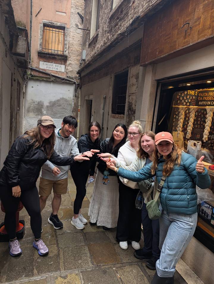 Group of smiling travellers in a narrow rain-wet Venetian alley extending their hands toward the camera.