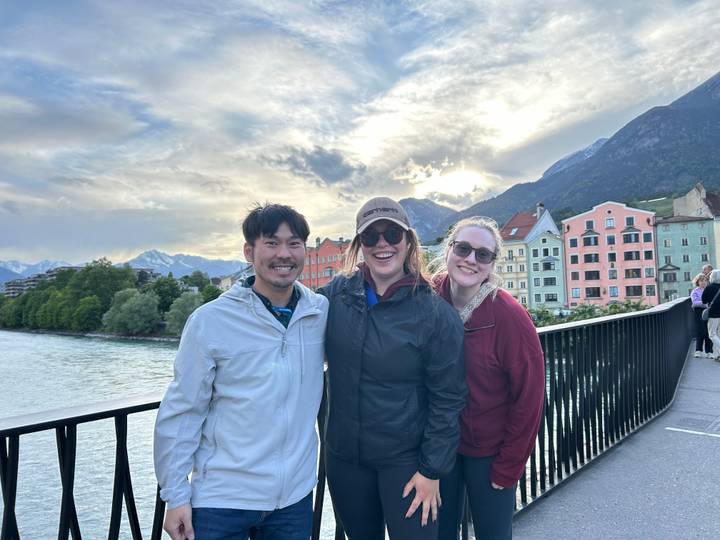 Three friends pose on a bridge with colourful riverside houses and snow-capped Alps in the background.