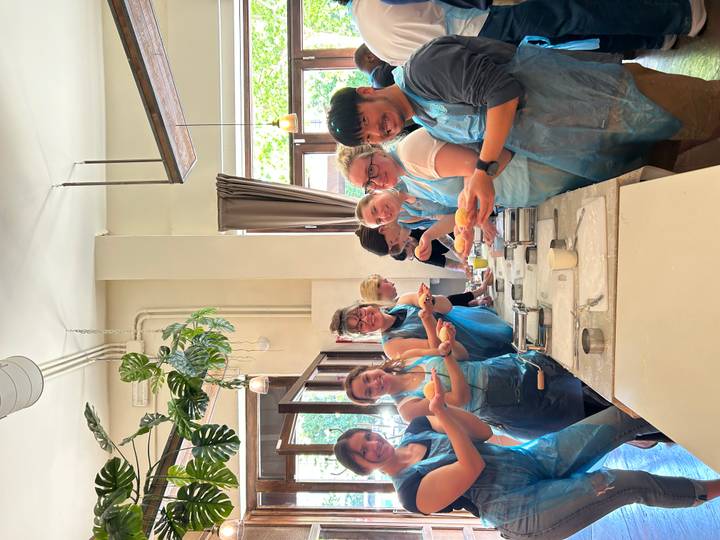 Cooking class scene with eight participants in blue aprons proudly holding freshly made pasta inside a bright studio.
