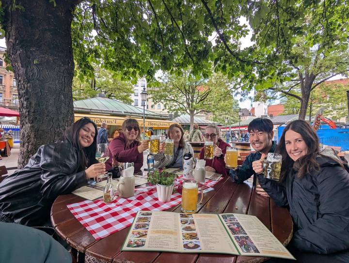 Friends raise large beer mugs and wine glasses at an outdoor Bavarian beer garden beneath leafy trees.